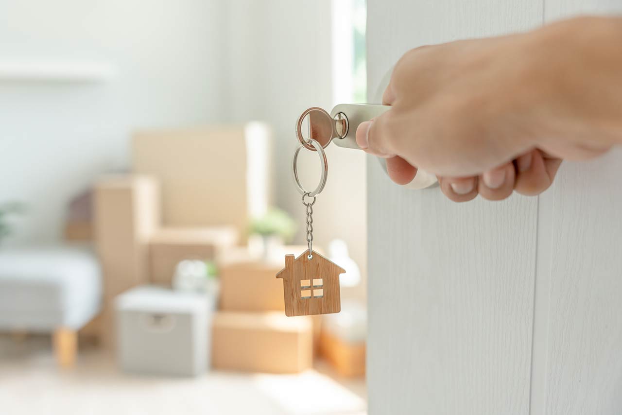 A key inserted into a door of the new house with a hand opening it, inside the room are cardboard boxes and furniture